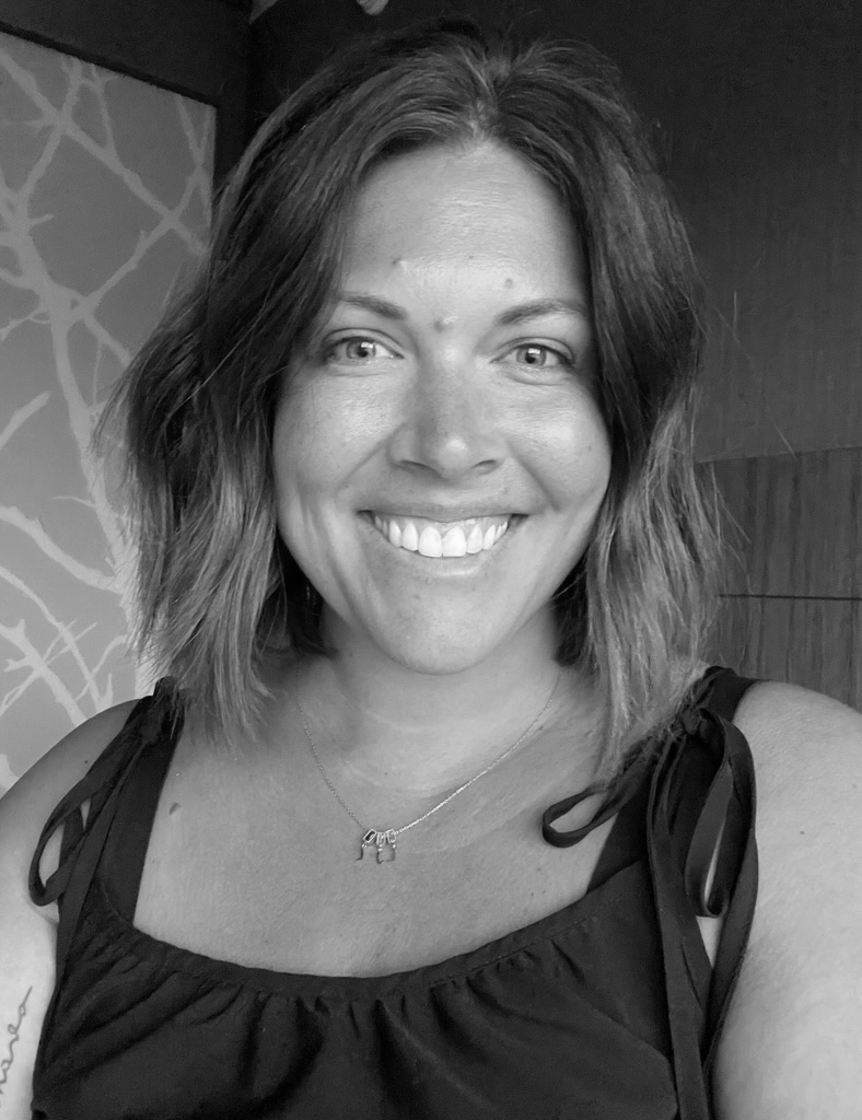Portrait of Morgan Ross, a woman with shoulder-length wavy hair, smiling brightly at the camera, wearing a dark sleeveless top with tie straps and a delicate necklace with small pendants, against a patterned wall background, in black and white