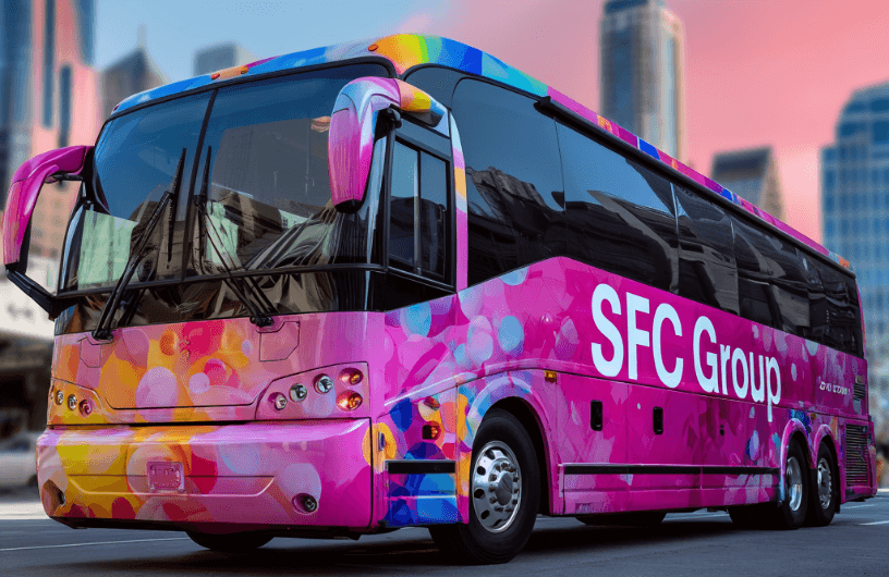 Colorful pink-dominant tour bus with vibrant multicolored bubble pattern wrap, "SFC Group" in large white letters on side, parked in city with tall buildings and pinkish sunset sky.